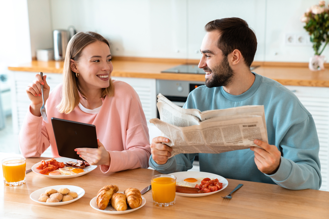 Ein junges Paar frühstückt in der Küche und liest dabei eine digitale und eine normale Zeitung.