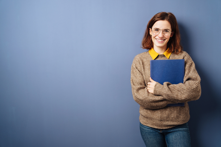 Eine Frau mit Brille steht vor einer blauen Wand und hält einen Ordner.