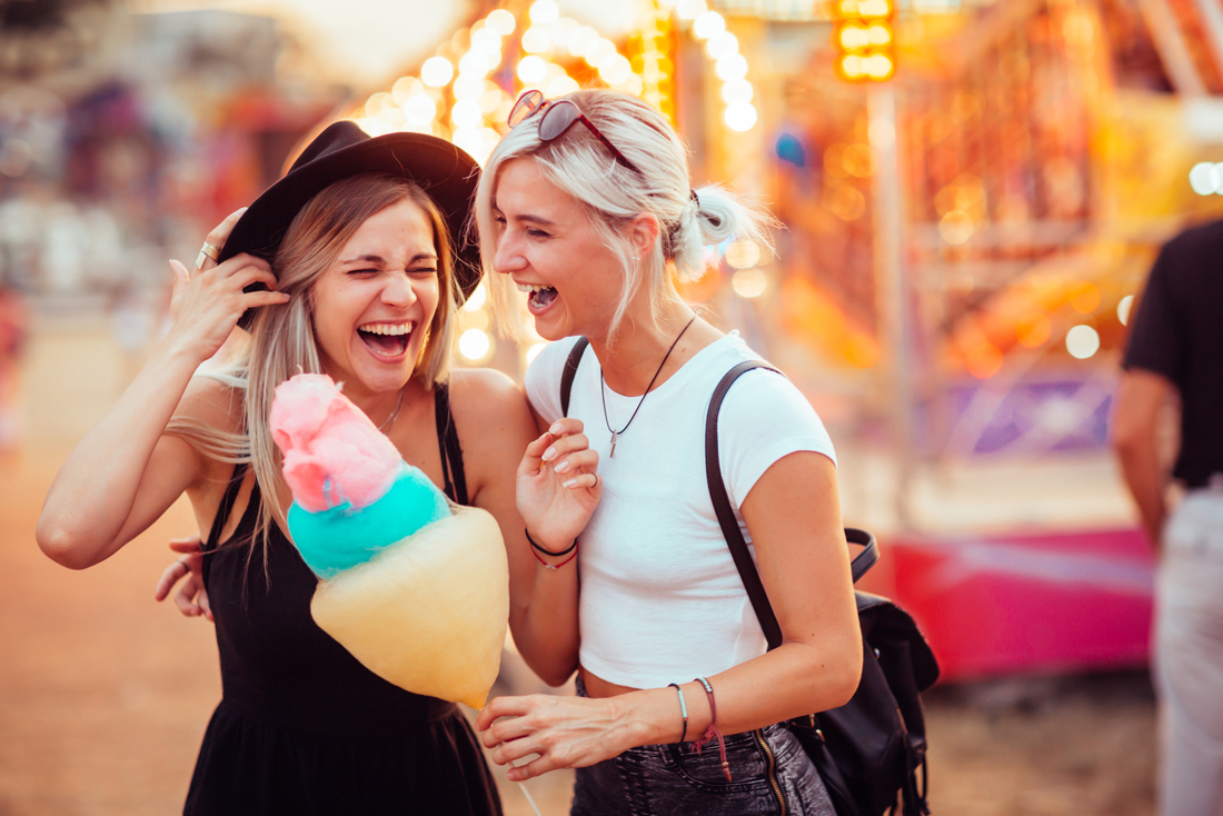 Zwei Frauen lachen fröhlich auf einem Jahrmarkt und halten Zuckerwatte in der Hand.