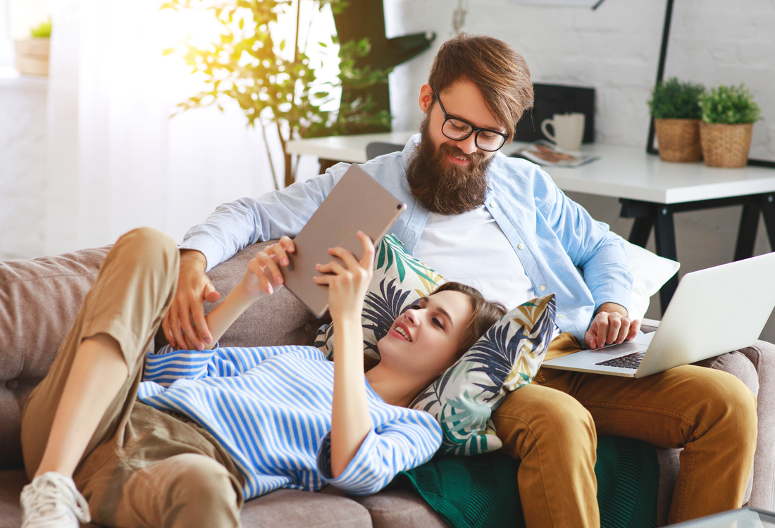 Ein Mann und eine Frau sitzen und liegen auf einem Sofa, der Mann arbeitet am Laptop, die Frau schaut auf ein Tablet.