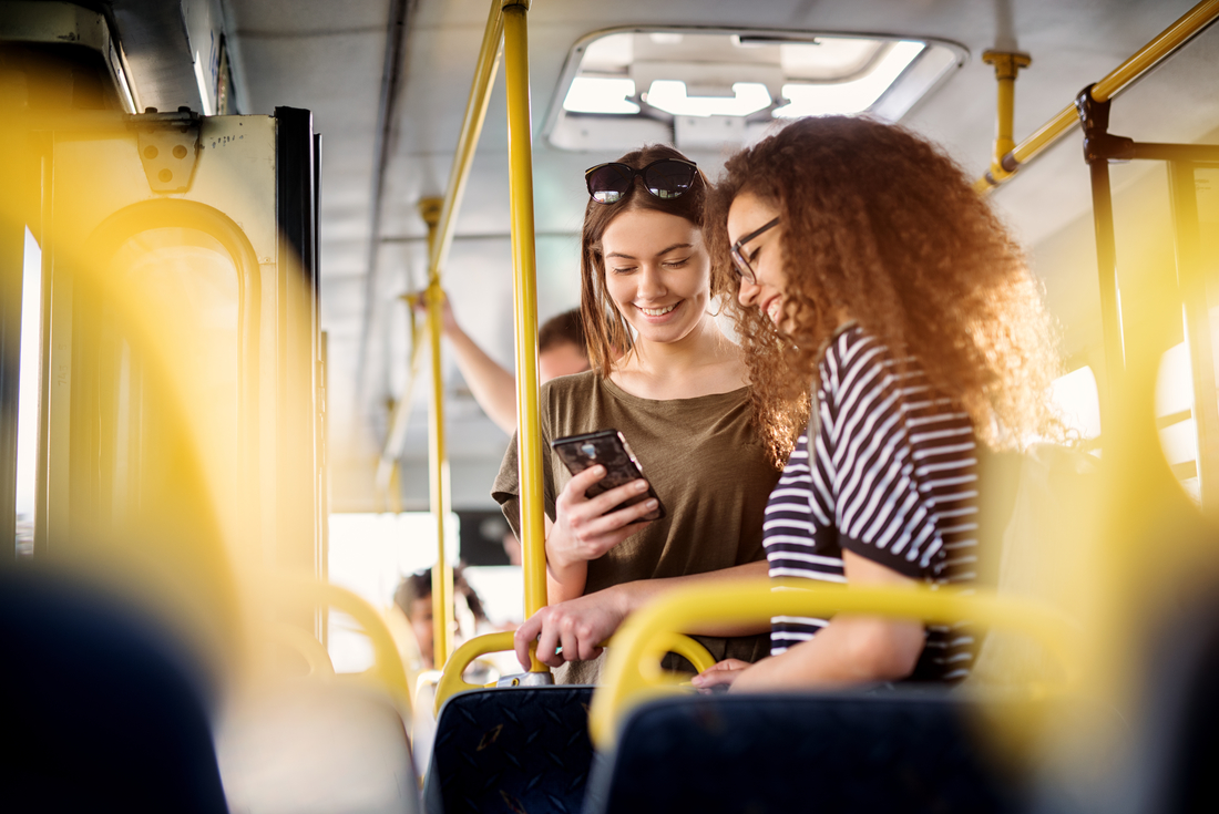 Zwei Frauen stehen im Bus, sie schauen auf ihre Handys und kommunizieren miteinander.