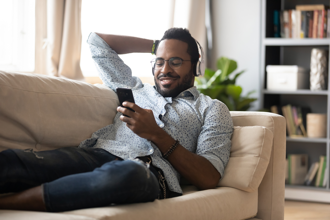Ein Mann liegt auf einer Couch und hält sein Handy in der Hand, er trägt Kopfhörer und lächelt.