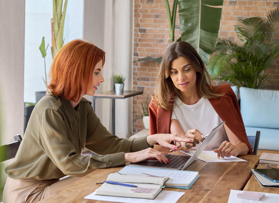 Zwei Frauen sitzen an einem Tisch und arbeiten gemeinsam an einem Laptop und Dokumenten.